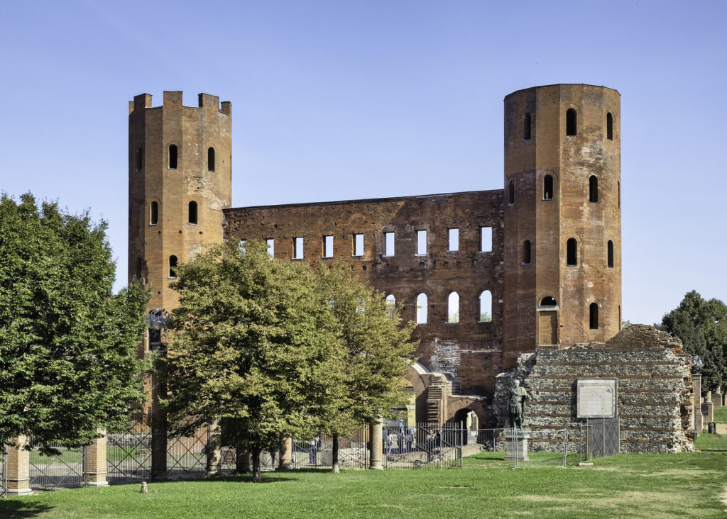 Frontansicht der Porta Palatina in Turin mit zwei hohen Backsteintürmen und der erhaltenen Mauerfront, umgeben von grünen Bäumen und Rasen.