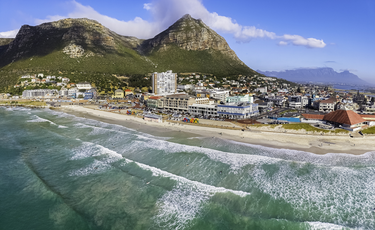 Luftaufnahme von Kapstadts Muizenberg-Strand mit bunten Badehäuschen, Wellen und Bergen im Hintergrund.