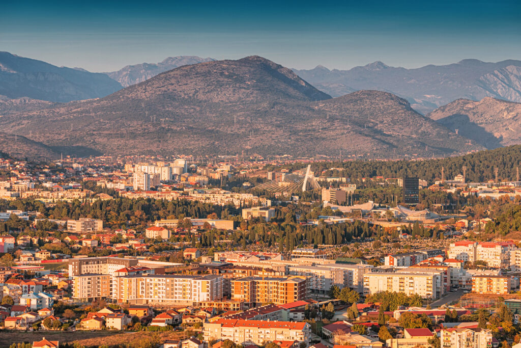 Panorama von Podgorica mit Wohnvierteln, viel Grün, der weißen Millennium-Brücke und den dahinter aufragenden Bergen in warmem Abendlicht.