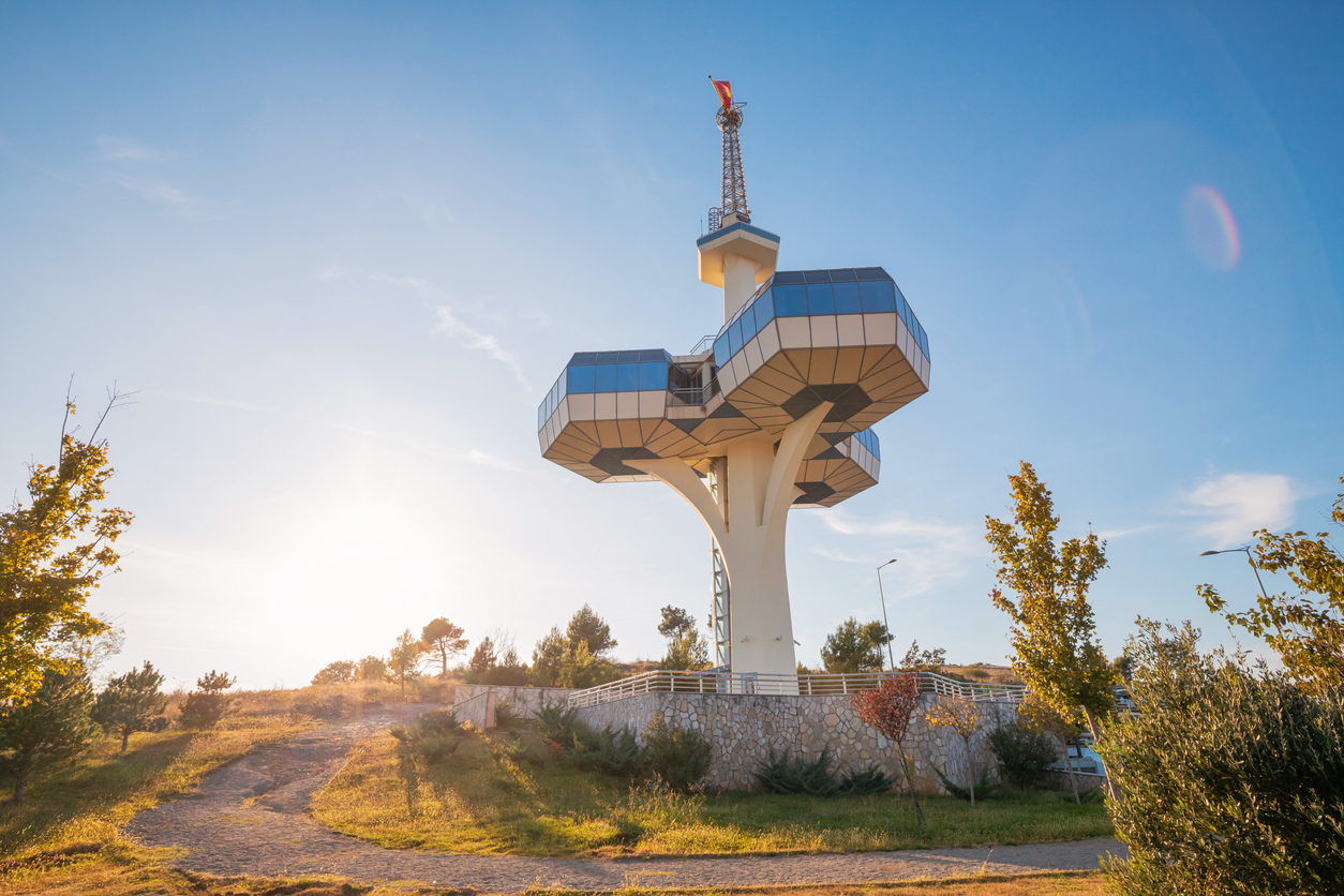 Futuristischer Aussichtsturm auf dem Hügel Dajbabska Gora bei Podgorica mit verglasten Plattformen, weißem Betonschaft und sonnigem Himmel; Grünflächen im Vordergrund.