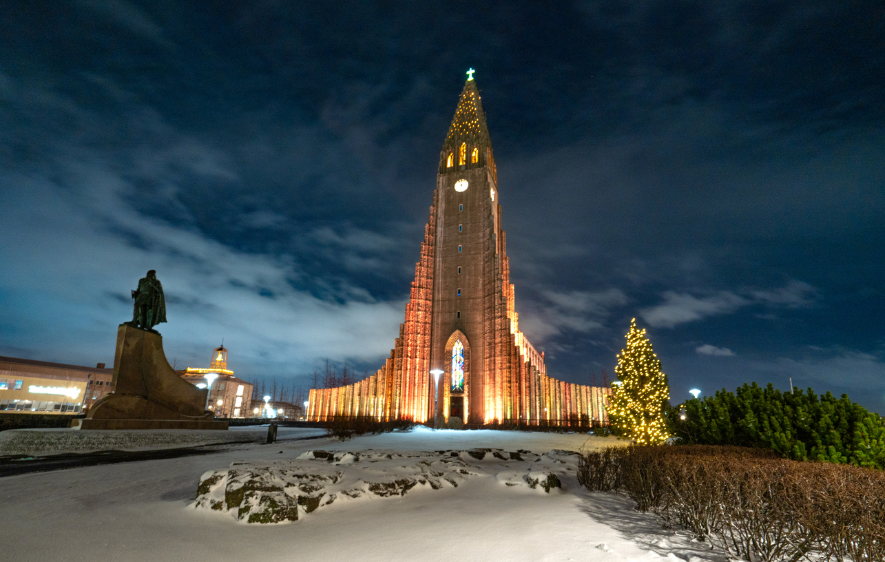 Beleuchtete Hallgrímskirkja in Reykjavík bei Nacht, verschneiter Platz mit Statue und Weihnachtsbaum.