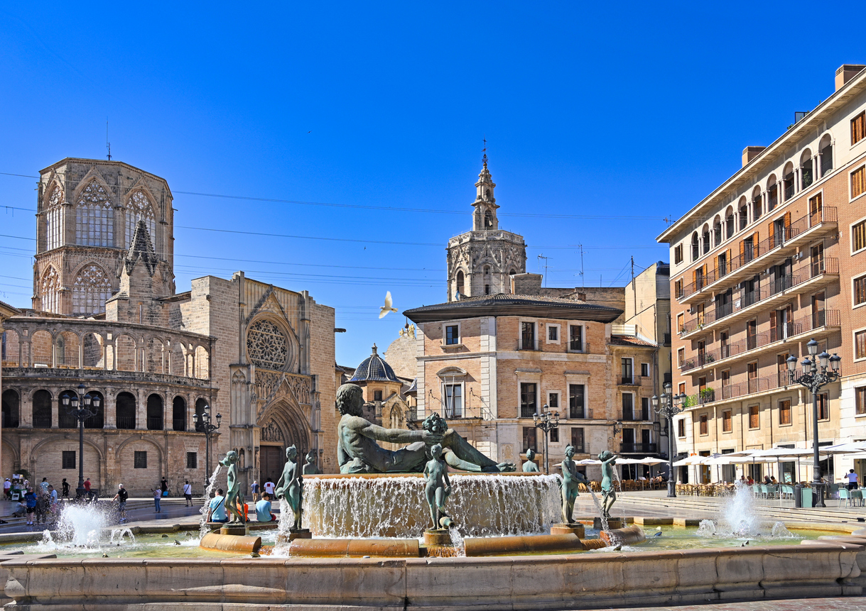 Plaza de la Virgen in Valencia mit dem Turia-Brunnen im Vordergrund, der gotischen Kathedrale und dem Miguelete-Turm bei klarem, blauem Himmel.