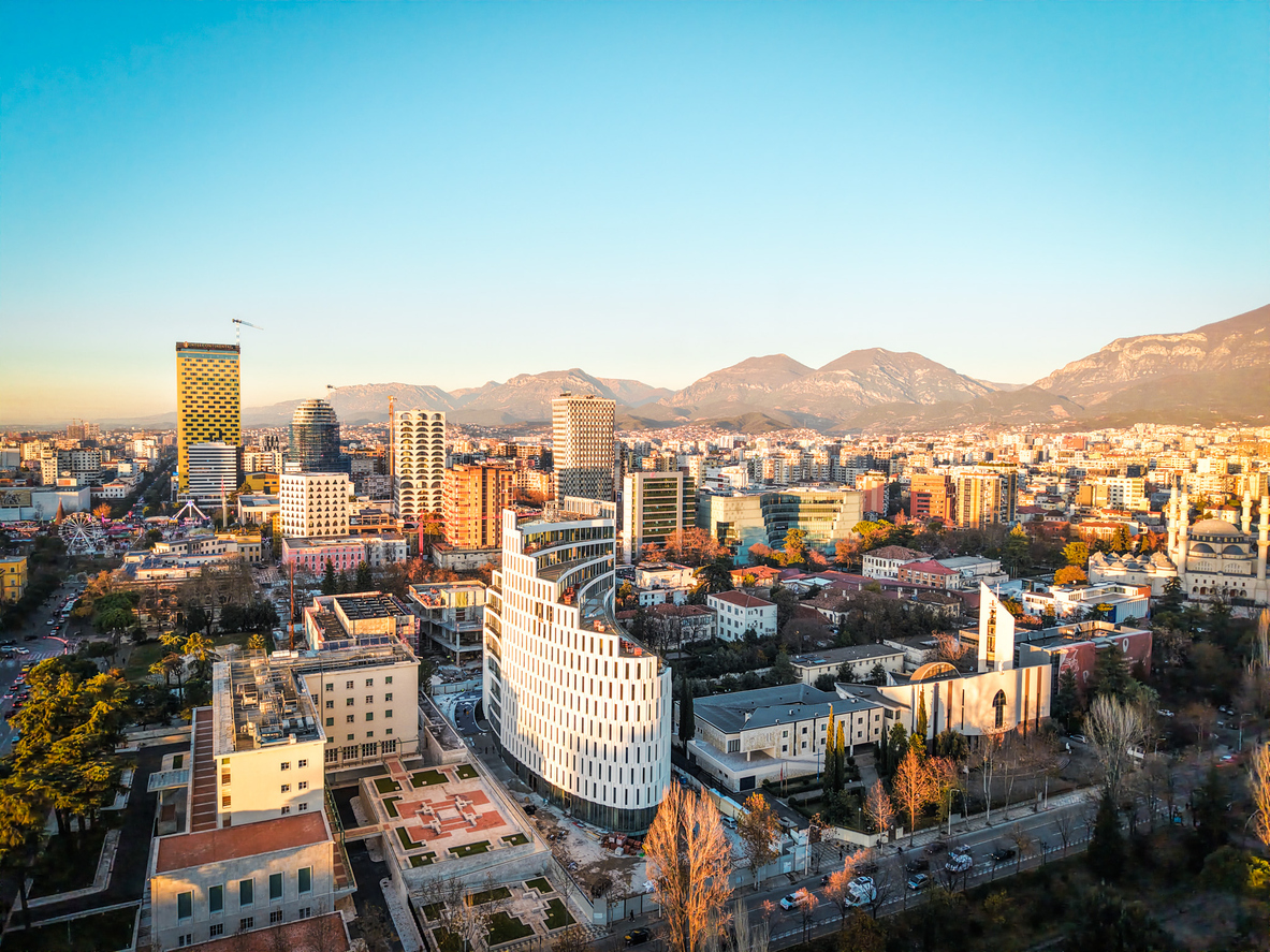 Luftaufnahme von Tirana mit Hochhäusern und modernen Neubauten im Stadtzentrum; im Hintergrund die Dajti-Berge, klarer Himmel und warmes Abendlicht