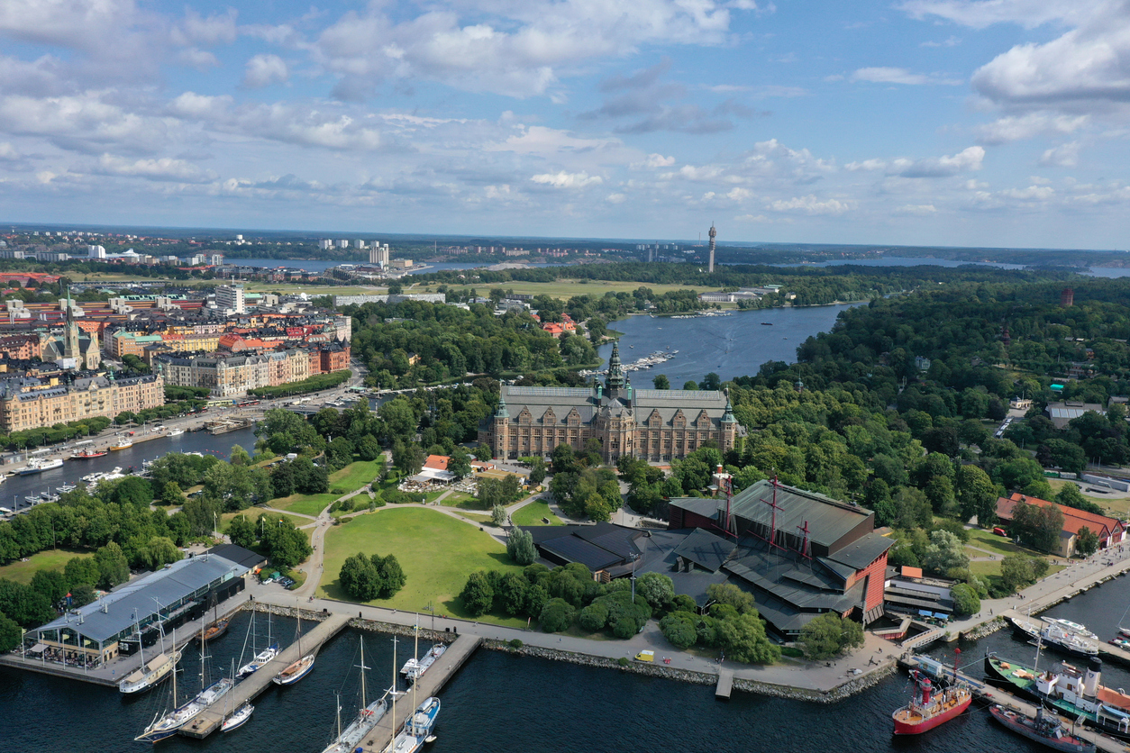 Stockholm, Luftaufnahme von Djurgården mit dem Nordiska museet im Zentrum, dem Vasamuseet rechts, Stege mit Segelbooten, weitläufige Grünflächen und Wasserläufe unter lockerem Wolkenhimmel