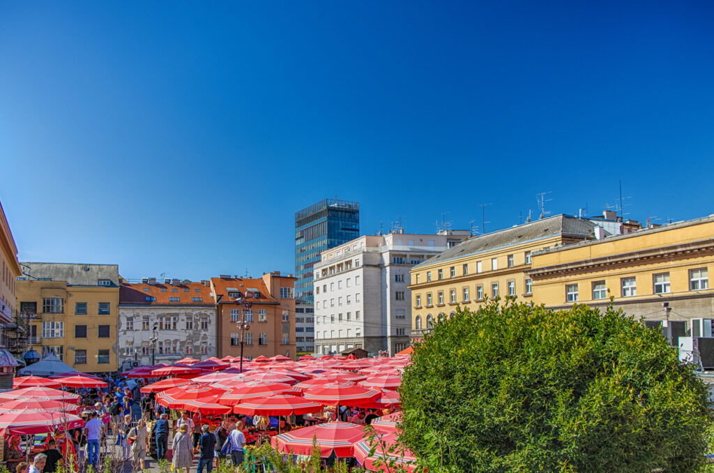 Blick auf den Dolac Markt in Zagreb mit zahlreichen roten Sonnenschirmen und Menschen zwischen historischen Gebäuden.
