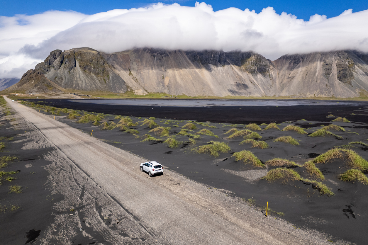 Auto auf isländischer Schotterstraße neben schwarzem Sand und Gras­dünen, Berge mit Wolkenhaube.