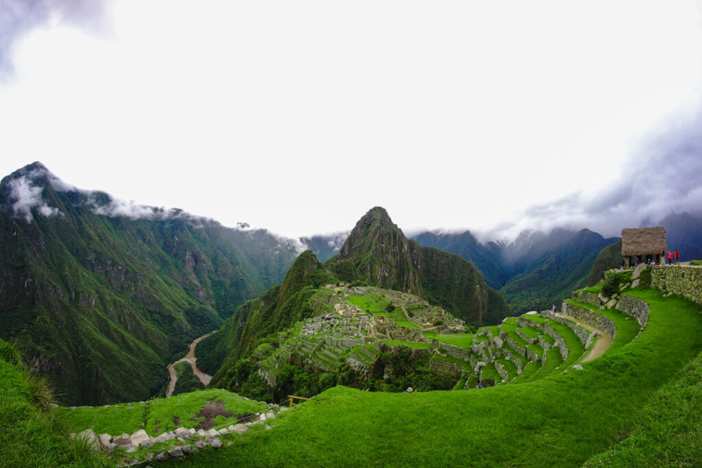 Panorama von Machu Picchu mit grünen Terrassen und Ruinen, Huayna Picchu dahinter; wolkenverhangenes Tal.