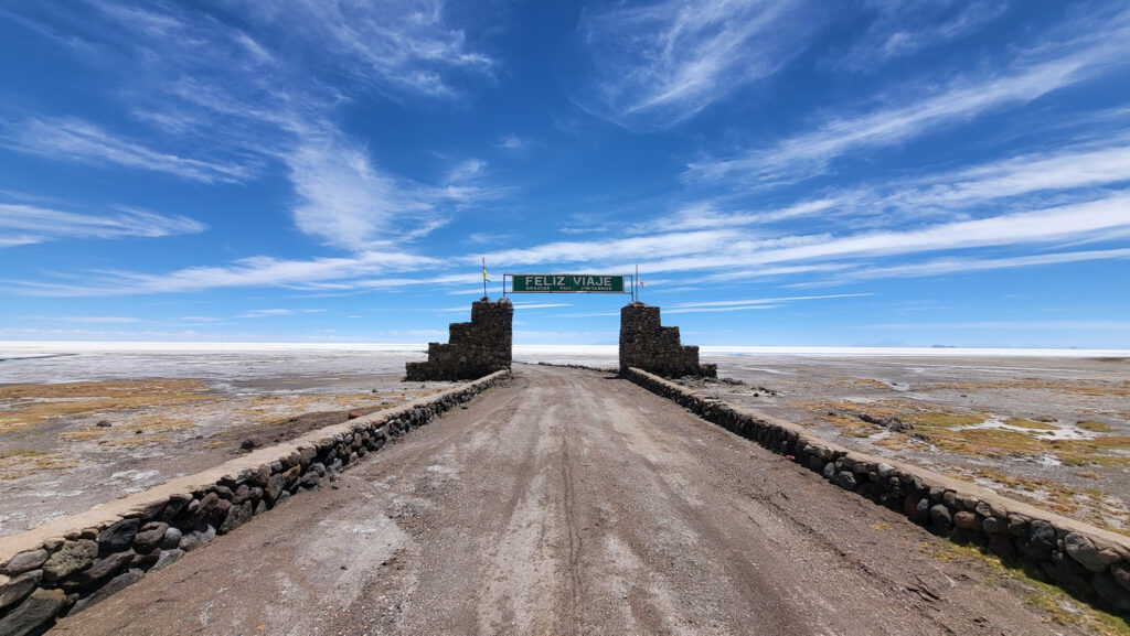 Steinportal mit „Feliz Viaje“-Schild über einer Piste, die in die weiße Salzwüste Uyuni führt; weiter blauer Himmel.