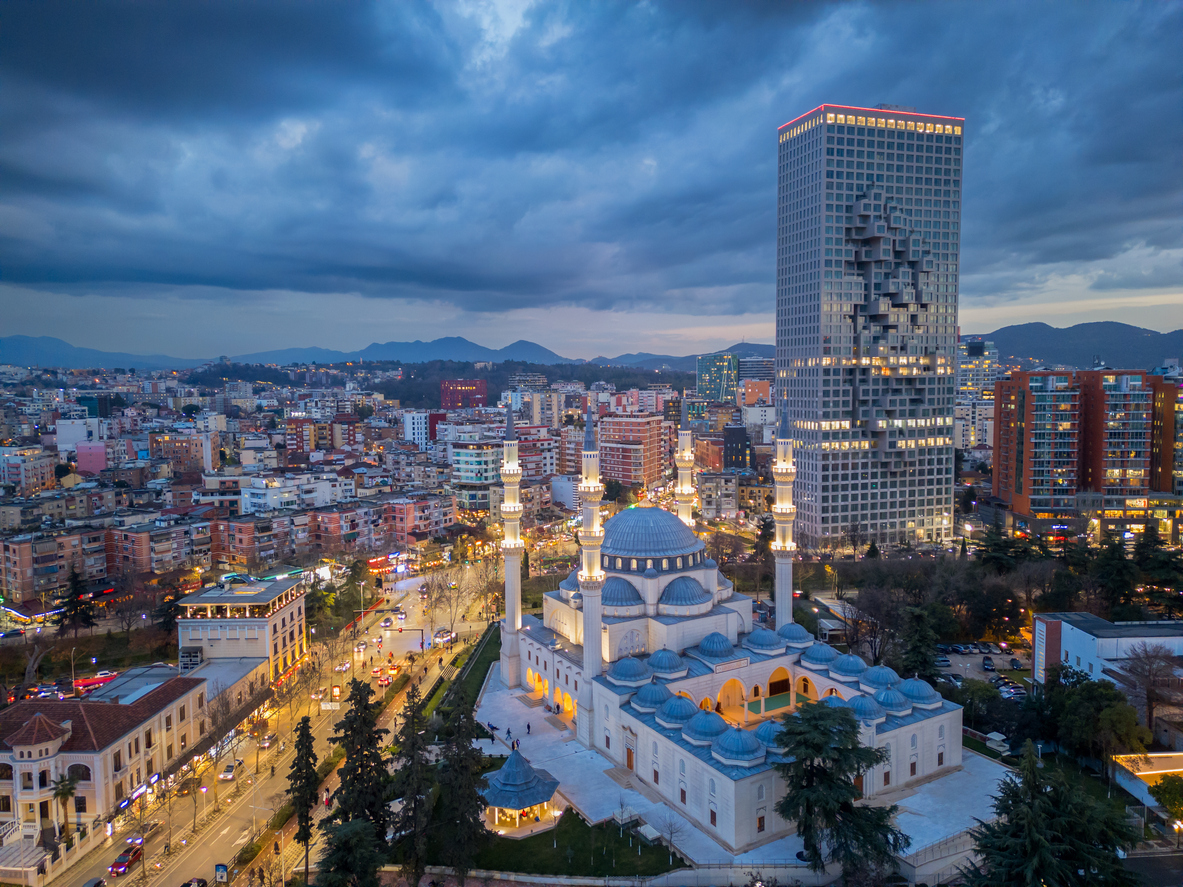Abendliche Luftaufnahme von Tirana mit der hell erleuchteten Großen Moschee und dem markanten Hochhaus „Downtown One“, umgeben von Stadtlichtern und Bergkulisse unter dramatischem Wolkenhimmel