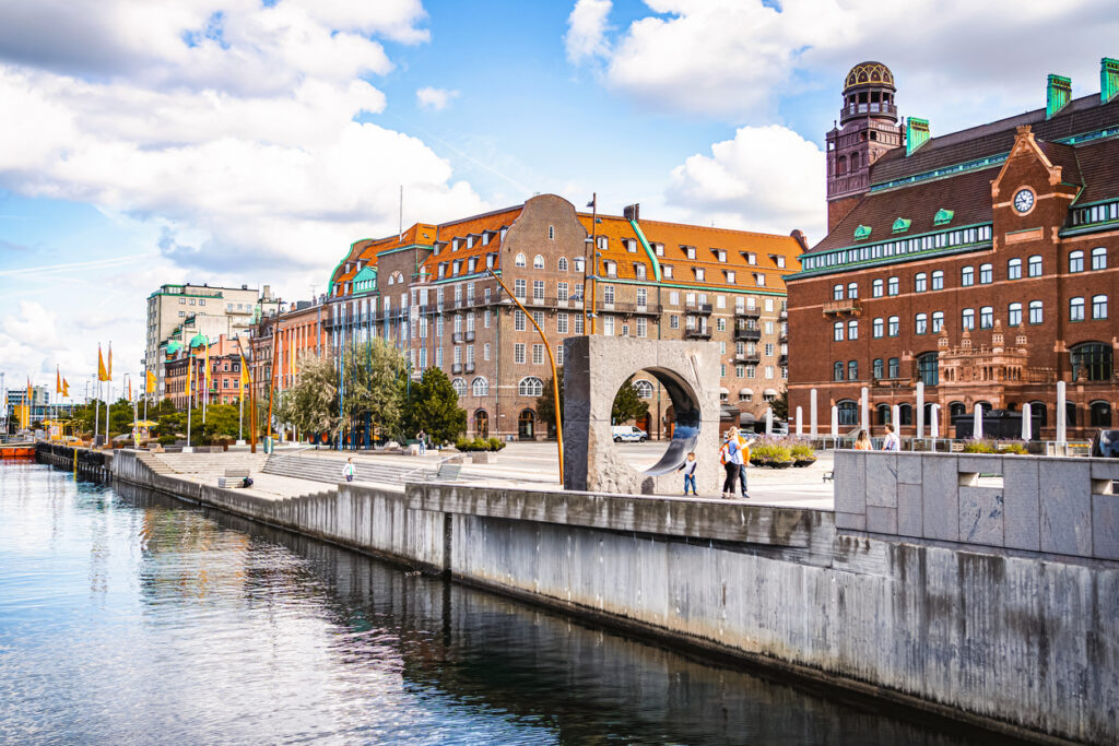 Uferpromenade in Malmö mit Backsteinbauten, rotem Ziegeldach, Uhrturm und moderner Skulptur am Wasser.