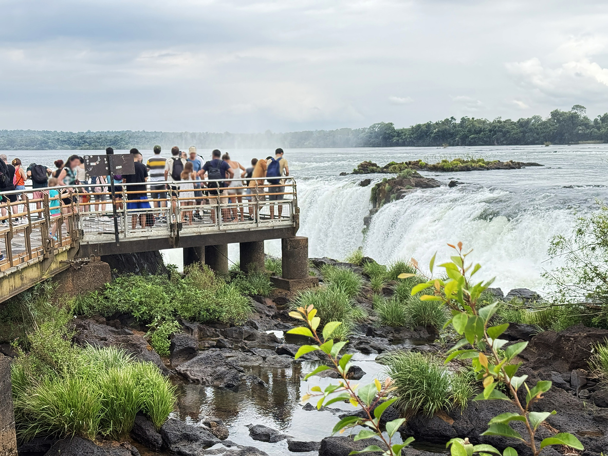 Besucher auf Metallsteg blicken auf breiten Wasserfall; Felsen, Grasinseln und bewölkter Himmel.