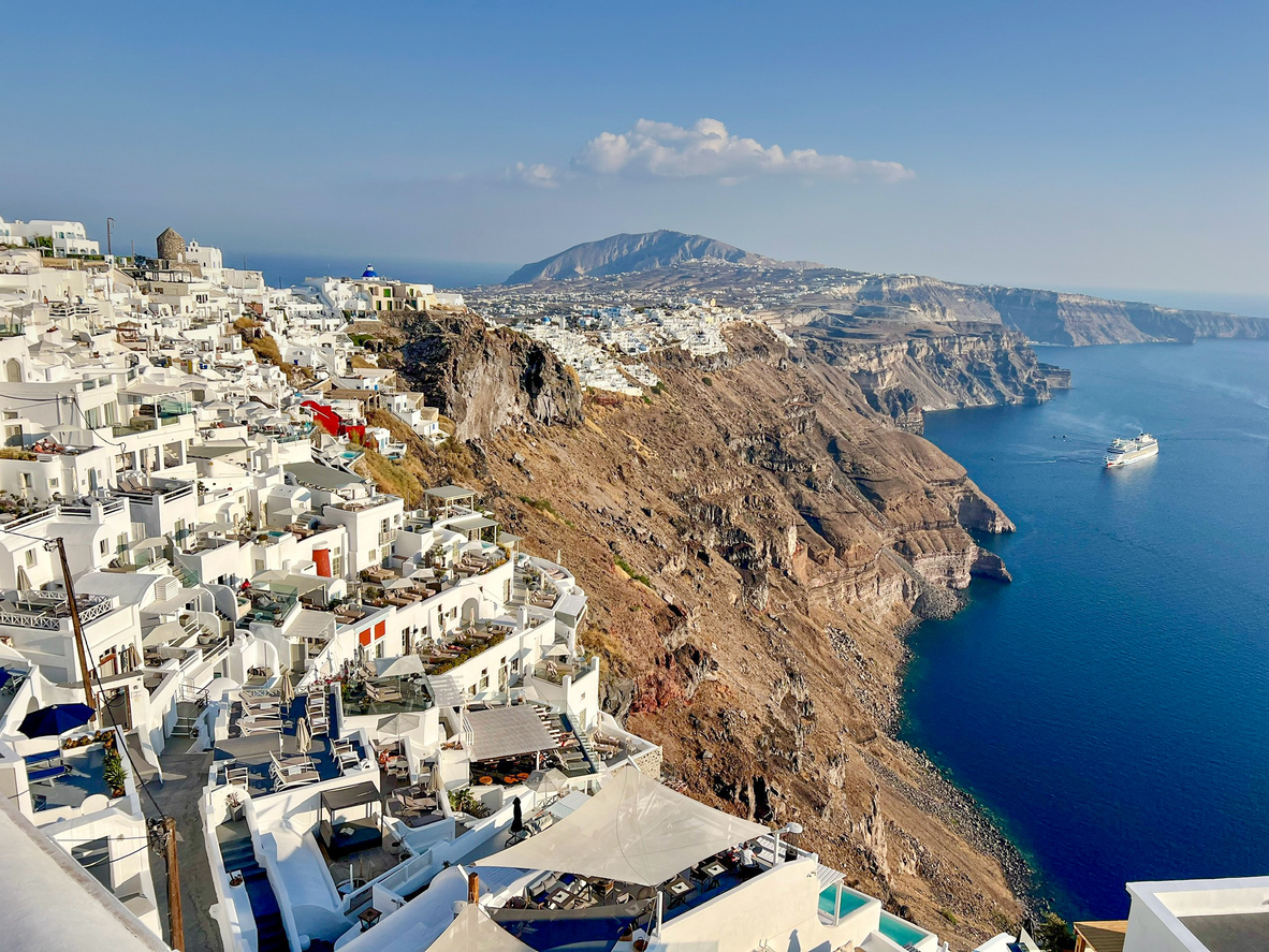 Weiße Häuser von Santorini auf Caldera-Klippen, darunter die blaue Ägäis mit Schiff, klarer Himmel.