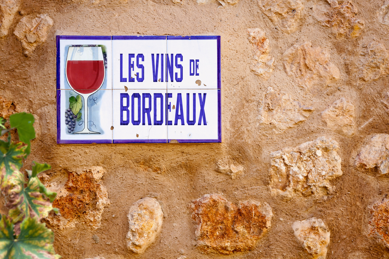 Nahaufnahme eines rechteckigen Keramikschildes mit violettem Rand und der Aufschrift „Les Vins de Bordeaux“, illustriert mit Weinglas und Trauben, befestigt auf einer ockerfarbenen, unregelmäßigen Steinwand; links ein Blatt im Vordergrund.
