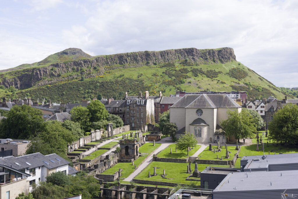 Edinburgh, Blick über einen historischen Friedhof mit Kirche und Grabmälern; dahinter die grünen Hänge von Arthur’s Seat und den Salisbury Crags unter leicht bewölktem Himmel