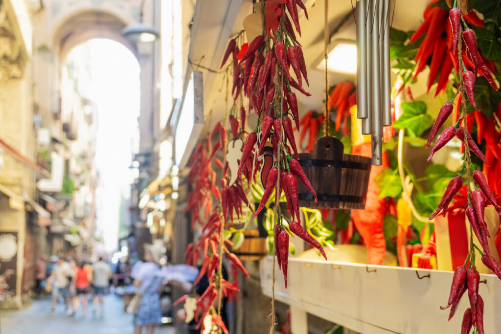 Neapel, schmale Altstadtgasse mit Marktständen; vorn hängen Bündel roter Peperoncini, unscharfe Passanten im Hintergrund, warmes Tageslicht