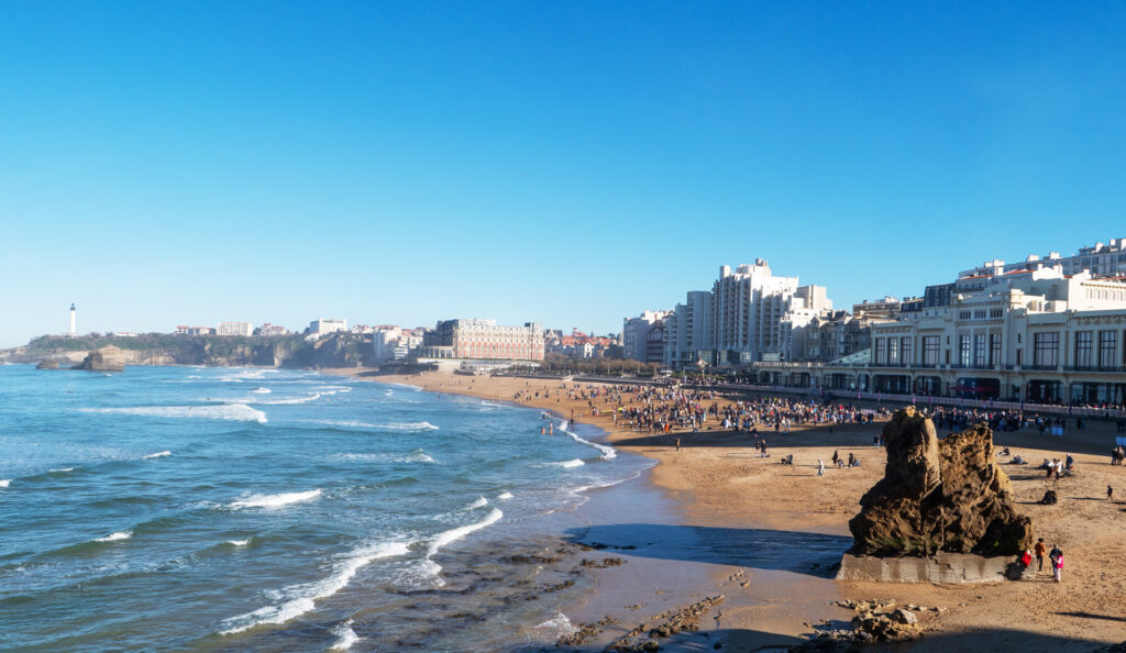 Weitläufiger Sandstrand von Biarritz in Frankreich mit vielen Menschen, die am Meer spazieren oder entspannen, im Hintergrund historische Gebäude und moderner Stadthintergrund.