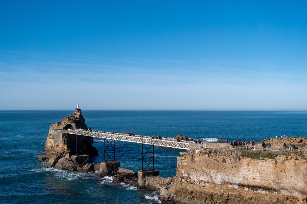 Küstenlandschaft von Biarritz mit dem bekannten Felsen „Rocher de la Vierge“, verbunden durch eine weiße Brücke mit dem Festland, im Hintergrund das weite Meer.