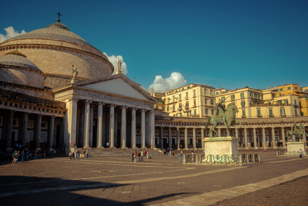 Neapel, Piazza del Plebiscito mit der Basilika San Francesco di Paola, Säulenportikus und großer Kuppel, Reiterstatue im Vordergrund, weiter Platz mit Passanten, warmes Abendlicht