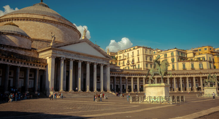 Neapel, Piazza del Plebiscito mit der Basilika San Francesco di Paola, Säulenportikus und großer Kuppel, Reiterstatue im Vordergrund, weiter Platz mit Passanten, warmes Abendlicht
