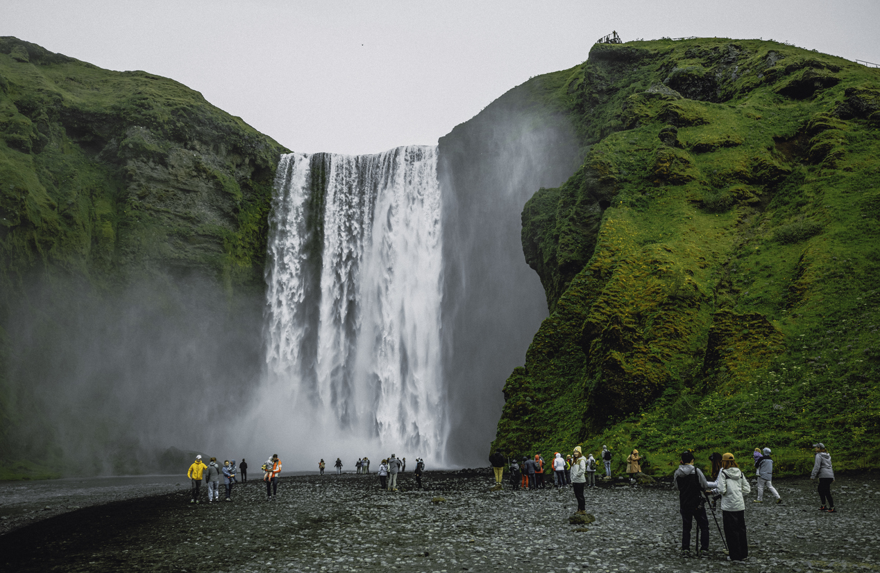 Skógafoss-Wasserfall mit Gischt, grünen Felsen und Touristen auf dem Kiesufer, Island.