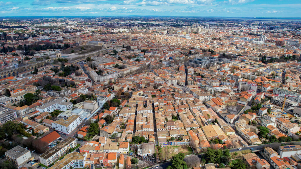 Weitwinklige Vogelperspektive auf Montpellier: dichtes Meer aus Terrakottadächern, links die langen Bögen des Aqueduc Saint-Clément und die Peyrou-Promenade mit Wasserreservoir; dahinter breitet sich die Stadt bis zum Horizont unter blauem Himmel aus.