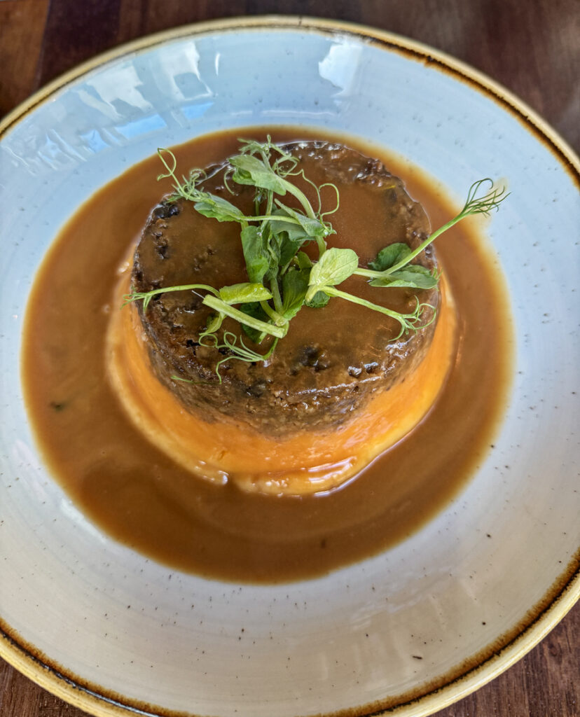 Runder Haggis auf orangem Püree, mit brauner Sauce übergossen und mit Erbsensprossen garniert, serviert auf weißem Teller.