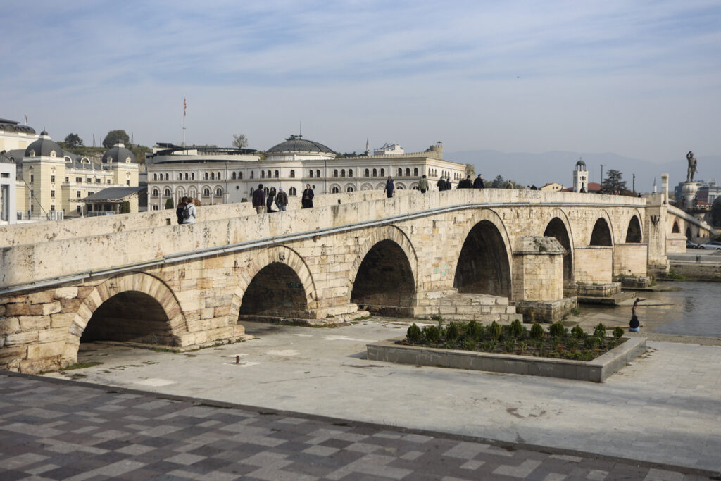 Historische Steinbrücke Kamen Most in Skopje mit mehreren Rundbögen über den Fluss Vardar; Fußgänger auf dem Brückenverlauf, dahinter neoklassizistische Museums- und Verwaltungsgebäude.
