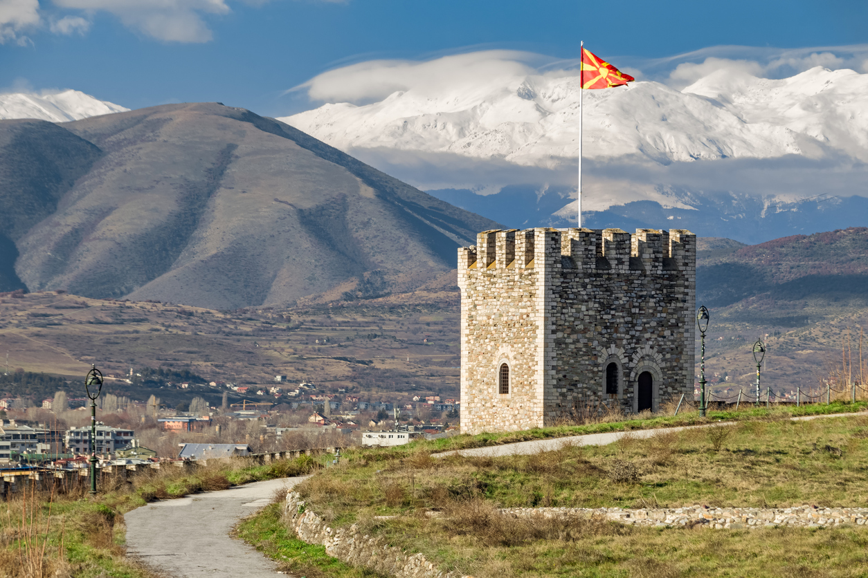 Steinturm der Kale-Festung in Skopje mit wehender Flagge Nordmazedoniens; im Hintergrund Hügellandschaft und schneebedeckte Berge unter wolkigem Himmel.