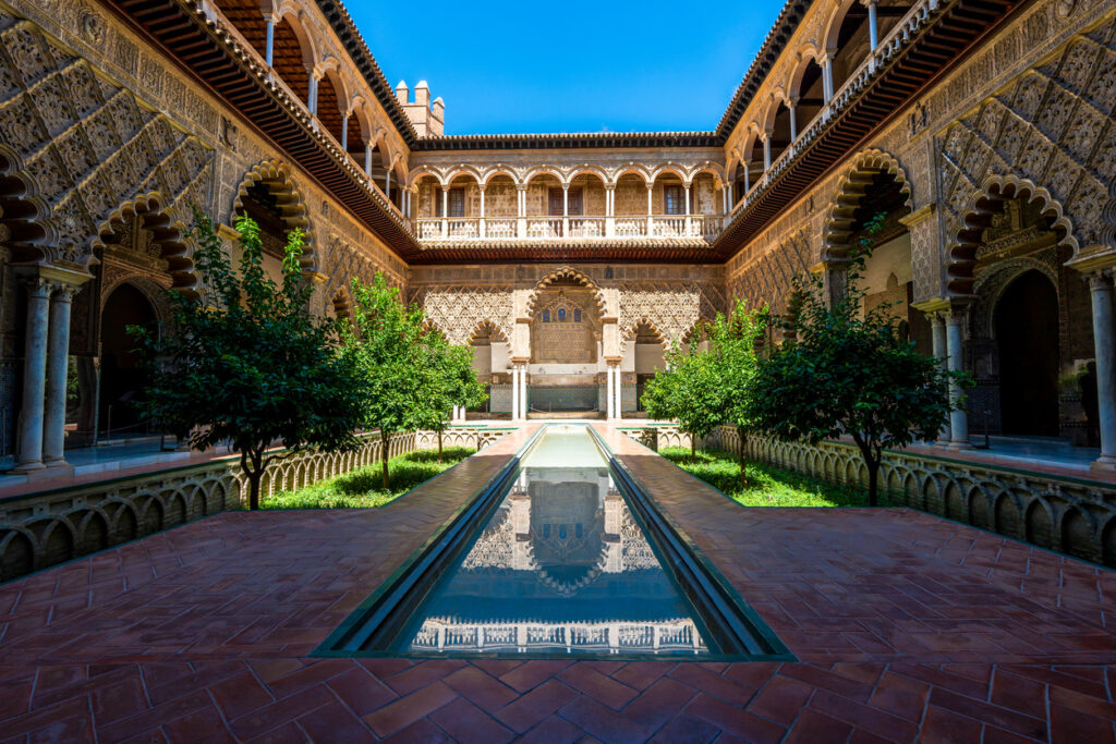Sevilla, Innenhof des Real Alcázar (Patio de las Doncellas) mit langem Wasserbecken, Orangenbäumen und reich verzierten Mudejar-Arkaden, symmetrische Ansicht bei Sonne