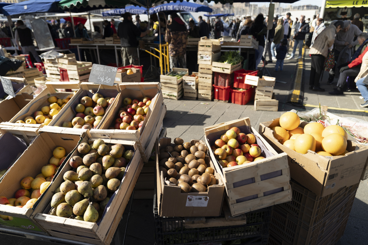 Holzkisten mit Äpfeln, Birnen, Kiwis und Grapefruits auf einem belebten Wochenmarkt am Vieux-Port in Marseille; Menschen und Marktstände im Hintergrund.
