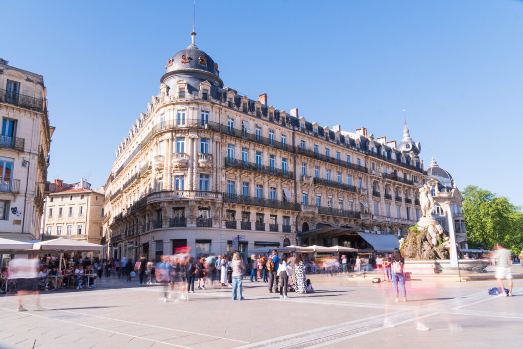 Belebter Platz in Montpellier mit eleganten Belle-Époque-Fassaden und Kuppeldach, Menschen in Bewegung und Brunnen mit Figurengruppe bei klarem blauem Himmel.