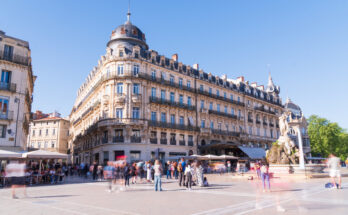 Belebter Platz in Montpellier mit eleganten Belle-Époque-Fassaden und Kuppeldach, Menschen in Bewegung und Brunnen mit Figurengruppe bei klarem blauem Himmel.