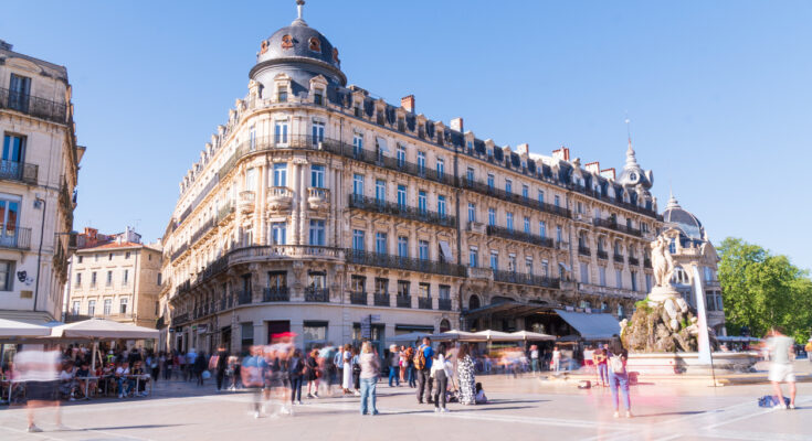 Belebter Platz in Montpellier mit eleganten Belle-Époque-Fassaden und Kuppeldach, Menschen in Bewegung und Brunnen mit Figurengruppe bei klarem blauem Himmel.