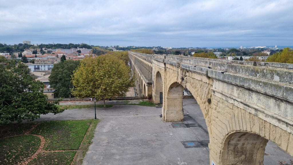 Historischer Kalkstein-Aquädukt mit langen, rhythmischen Bögen, der sich perspektivisch in die Ferne zieht; darunter ein Platz mit Bäumen, links Dächer der Stadt, bedeckter Himmel.