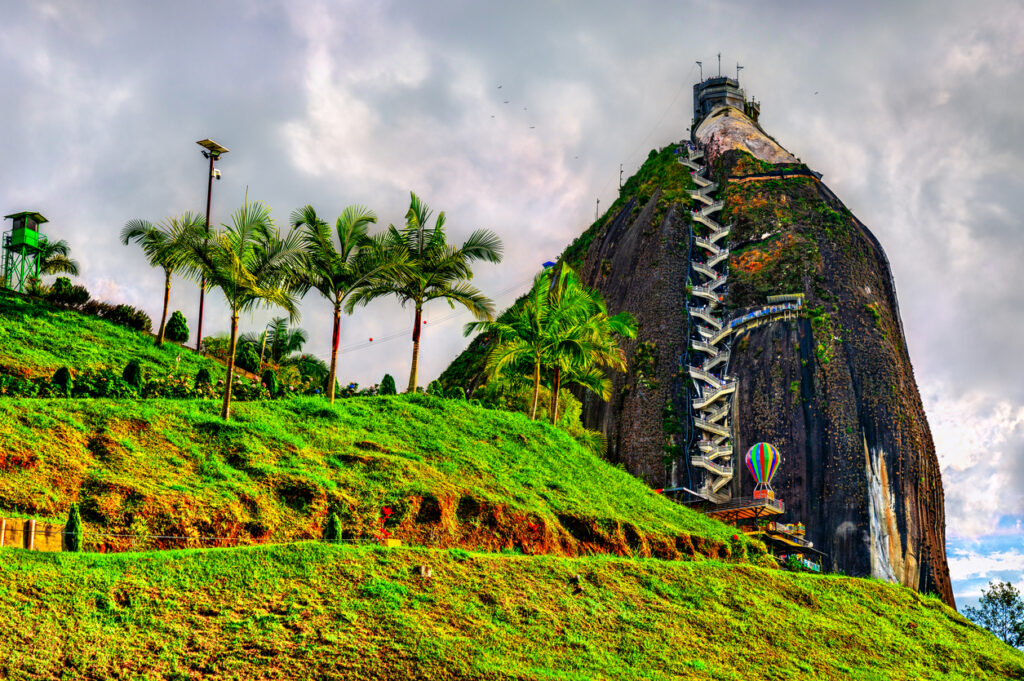 Der Felsen von Guatapé (La Piedra del Peñol) in Antioquia, Kolumbien, ein gewaltiger Monolith mit markanter Außentreppe, leuchtend grünen Hügeln und Palmen unter dramatischem Himmel.