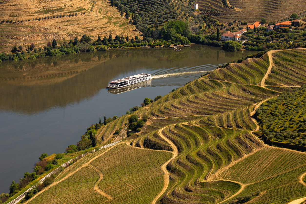 Flusskreuzfahrtschiff gleitet über den ruhigen Douro, rechts und links geschwungene Terrassenweinberge mit Wegen und Quintas; warme Nachmittagsstimmung, klare Sicht.