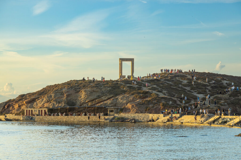 Portara auf Naxos mit vielen Besuchern auf Hügel, Uferpromenade am Meer, warmer Abendhimmel.