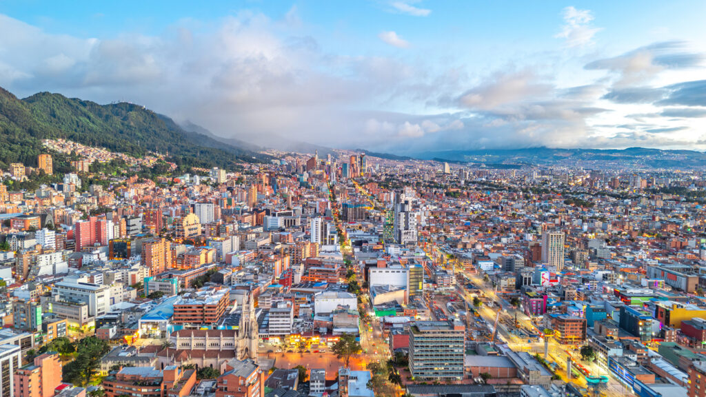 Panorama von Bogotá, Kolumbien, mit dichter urbaner Bebauung, Hochhäusern, Kirchen, beleuchteten Straßen und den grünen Anden im Hintergrund unter wolkenverhangenem Himmel.