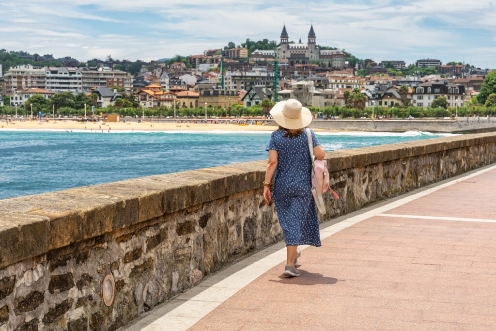 Frau mit Sommerhut und Rucksack spaziert an der Promenade entlang der La Concha in San Sebastián mit Blick auf Strand, Meer und Stadtpanorama.