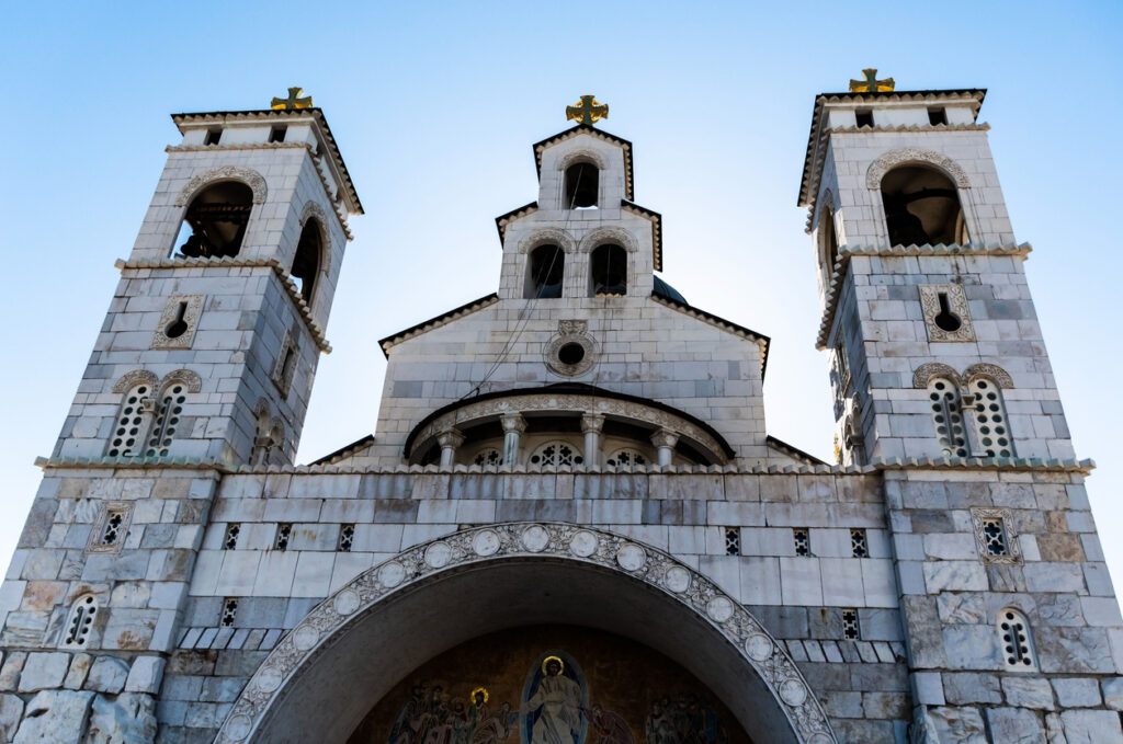 Untersicht der orthodoxen Kathedrale der Auferstehung Christi in Podgorica mit zwei weißen Glockentürmen, vergoldeten Kreuzen und blauem Himmel im Gegenlicht.
