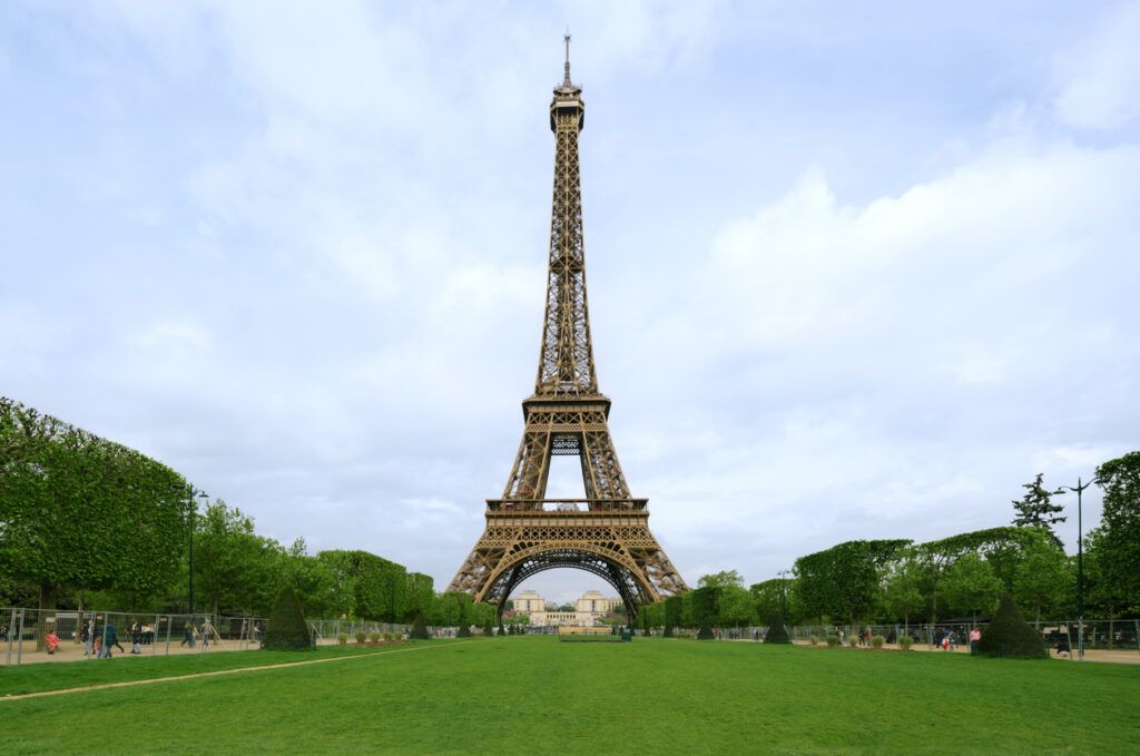 Blick auf den Eiffelturm in Paris, Frankreich, mit grünen Rasenflächen und Bäumen im Vordergrund unter bewölktem Himmel.