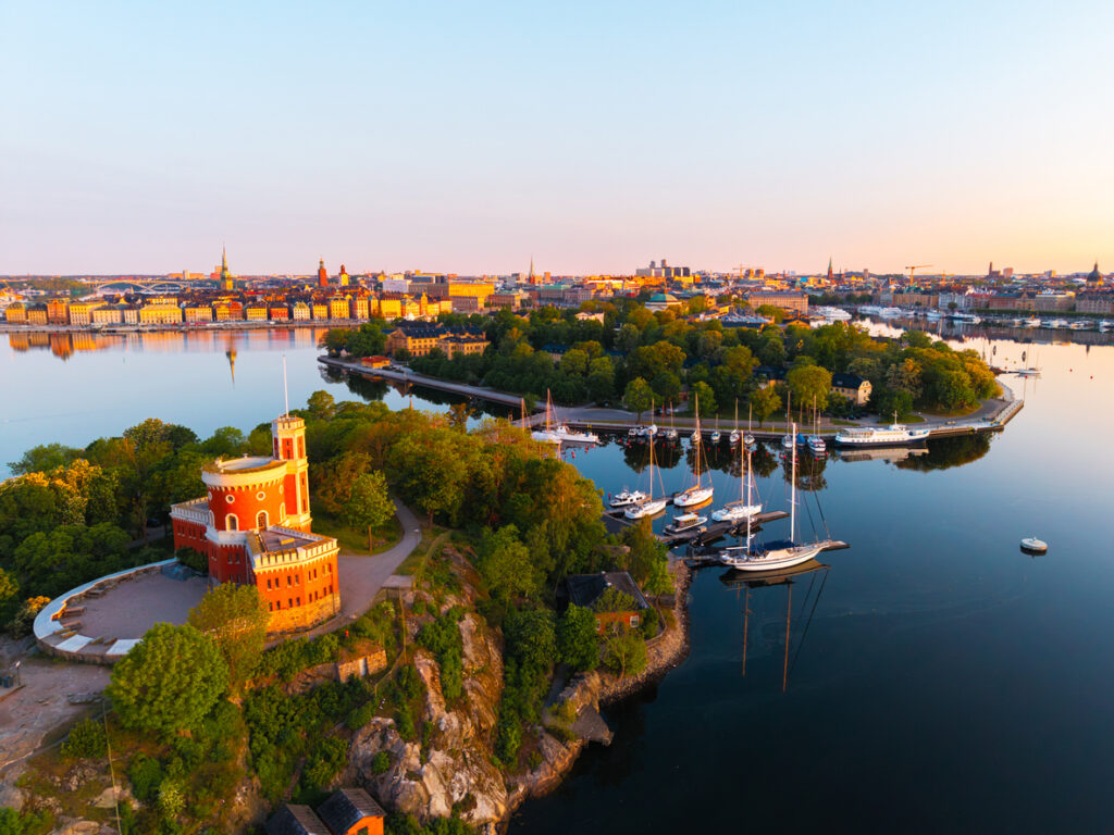 Stockholm, Luftaufnahme von Kastellholmen mit dem roten Kastellet auf Felsen, Segelboote an der Marina, ruhige Wasserfläche mit Spiegelungen, warme Abendstimmung