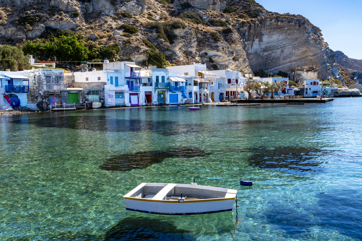 Fischerhäuser auf Milos am Ufer mit kleinem Boot im türkisblauen Wasser vor Felsklippen.