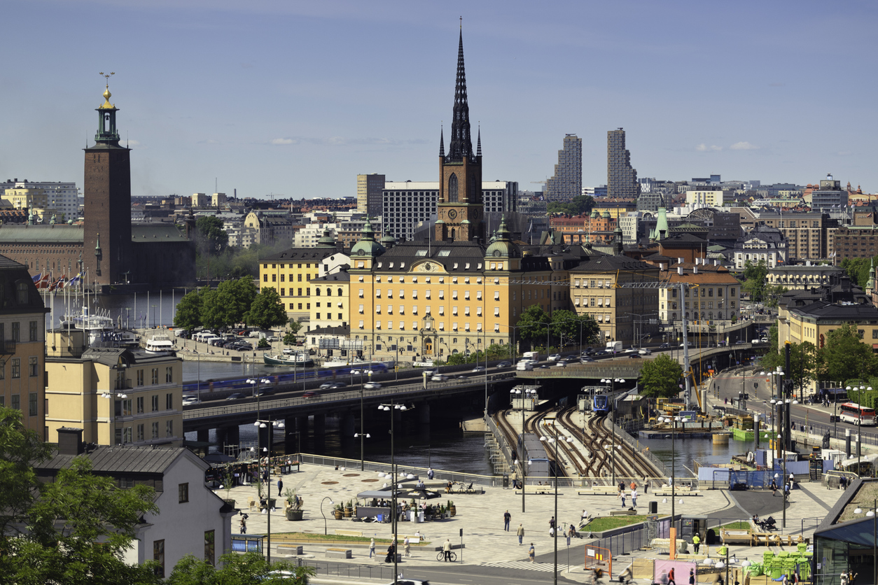 Stockholm, Stadtansicht mit Riddarholmskyrkan im Zentrum und dem Stadshuset links am Wasser, Brückenverkehr, U-Bahn-Gleise und Platz im Vordergrund, sonniger Tag