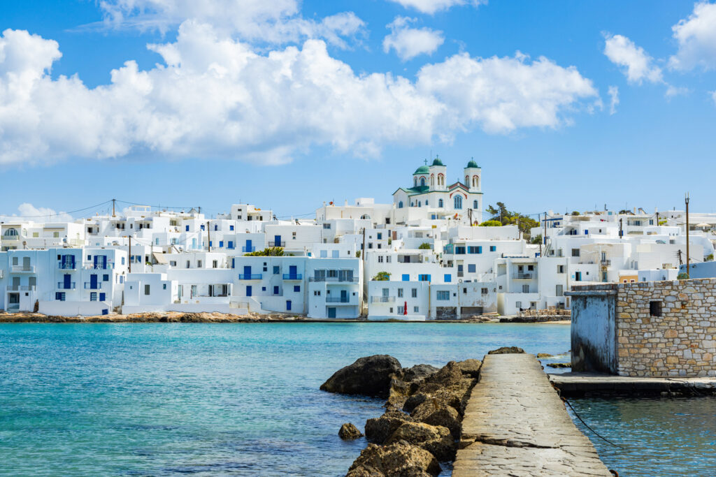 Weiß-blaue Häuser und Kirche auf Paros am Meer, Steinpier im Vordergrund, blauer Himmel.