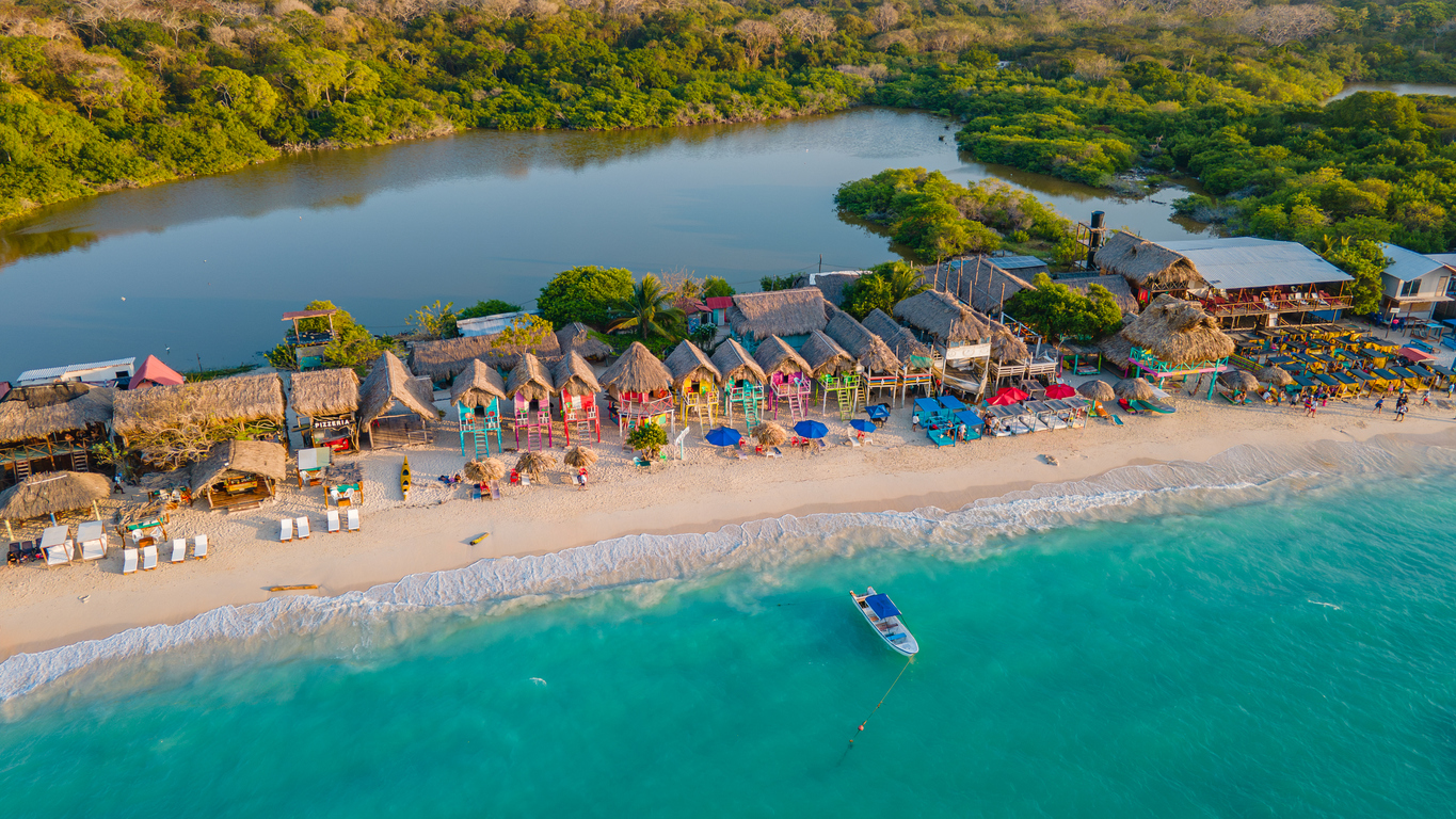 Luftaufnahme von Playa Blanca in Cartagena, Kolumbien, mit türkisblauem Meer, weißem Sandstrand, bunten Hütten mit Strohdächern und üppiger Vegetation im Hintergrund.