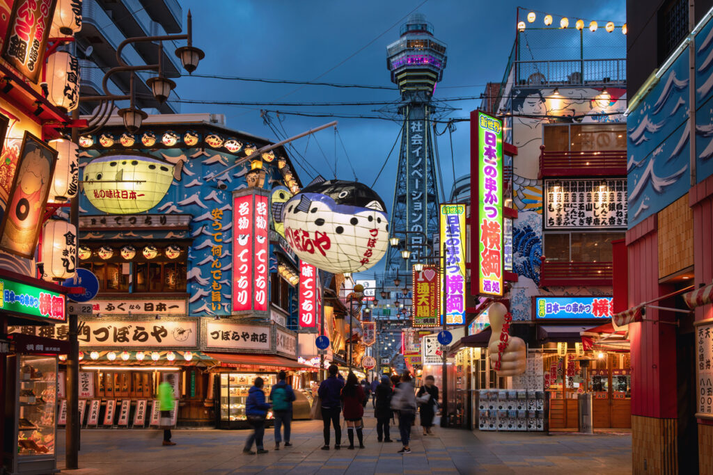 Abendstimmung in Osaka mit Menschen, die durch das bunte Viertel Shinsekai spazieren, leuchtenden Reklametafeln und dem Tsutenkaku-Turm im Hintergrund.