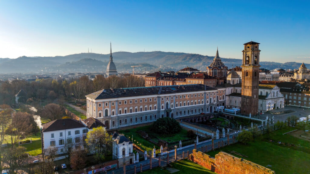Panorama von Turin bei Morgenlicht mit dem markanten Turm der Mole Antonelliana, dem Glockenturm des Doms, barocken Kuppeln und dem langgestreckten Palazzo Reale; im Hintergrund die grünen Hügel der Stadt.