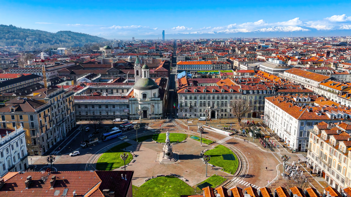 Luftaufnahme über das Zentrum von Turin mit einer kreisförmigen Piazza samt Statue, roten Ziegeldächern, einem geradlinigen Boulevard und dem Intesa-Sanpaolo-Tower; im Hintergrund schneebedeckte Alpen.