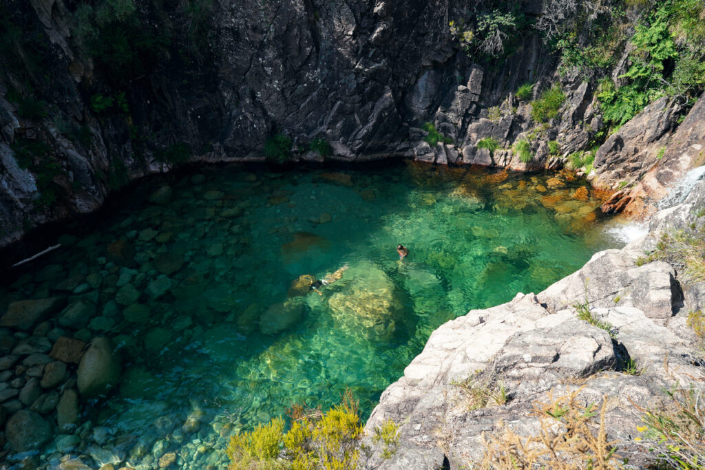 Naturpool mit türkisgrünem Wasser, Felsen und zwei Badenden, kleiner Zufluss am Rand.